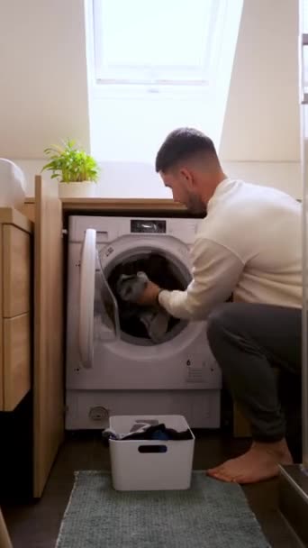 男性は洗濯機 洗濯室の主要な電化製品 ハードウッドフローリング 近くのキッチンにガソリンストーブに服を入れています — ストック動画
