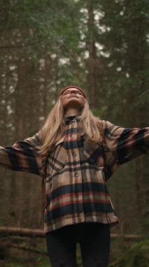 Young woman making deep breath and enjoying the fresh air while walking in the misty forest on fall day. Lady raised arms, closed eyes around amazing nature