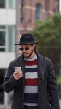 Paltolu ve güneş gözlüklü yakışıklı esmer adam dışarıda gündüz dolaşırken akıllı telefon kullanıyor. Akıllı adam taksi çağırmak için başvuru bekliyor.