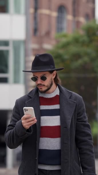 夏時間の屋外散歩中にスマートフォンを使用してコートやサングラスで幸せなハンサムなブルネットの男 アプリケーションによるタクシー注文を待っているスマート男 — ストック動画