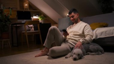 Yaşam tarzı konsepti. Akıllı telefonu kullanan gülümseyen bir adamın portresi. Halıda köpeğiyle oturuyor ve onu okşuyor. Adam evde vakit geçiriyor, sosyal medyada sohbet ediyor, mesaj yazıyor ya da mesaj okuyor.