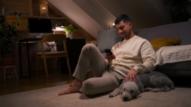 ライフスタイルコンセプト 犬と一緒にカーペットの上に座っているスマートフォンを使用して笑顔の男の肖像画 自宅で時間を過ごしたり ソーシャルメディアでチャットしたり Smsをテキストしたり メッセージを読んだりする 動画クリップ