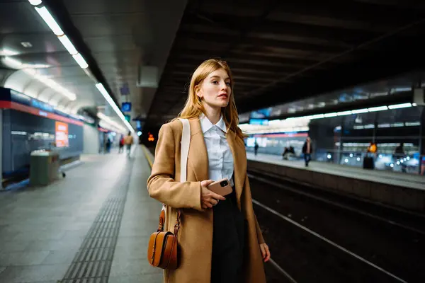 地下鉄のスマートフォンでワイヤレスインターネットをサーフィンするスーツの真剣なビジネスマン ソーシャルメディアと待合列車を使用して 携帯電話で地下鉄プラットフォームに立っている女性 ロイヤリティフリーのストック写真