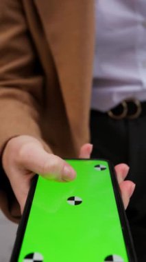 Sokakta dikey olarak yeşil ekran tutan ve telefona dokunan genç bir kadının dikey görüntüsü. İnternet teknolojisi kavramı