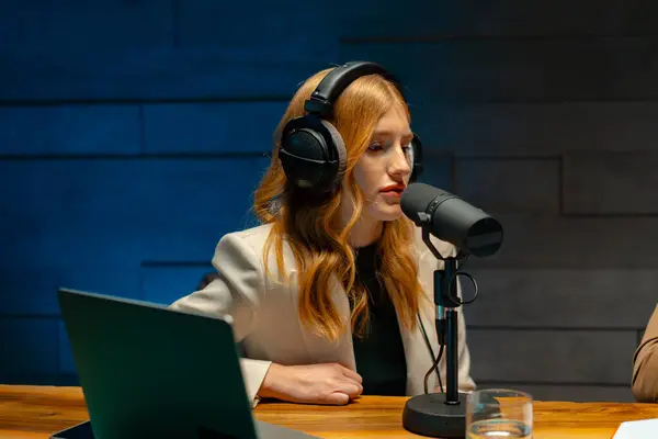 ヘッドフォンを着用している女性は イベントで話す準備ができて マイクの前に机に座っています 彼女は ラップトップに接続されたオーディオ機器を使用して スポークスマンです ストック画像