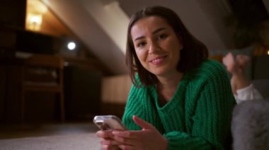 Genç bir kadın oturma odasında yere uzanmış ve mutlu yüzlü bir akıllı telefon kullanıyor. Kameraya bakan hoş bir bayan.