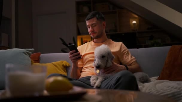 彼の部屋で携帯電話を使用している若い男は 夜にリラックスして彼の犬と抱き合って — ストック動画