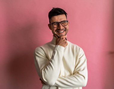 Gözlüklü ve beyaz kazaklı bir adam pembe bir duvarın önünde mutlu bir şekilde gülümsüyor. Görme yeteneği sakalını ve çene hattını tamamlıyor.