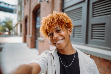 Kızıl saçlı ve kâküllü mutlu genç bir adam binanın önünde selfie çekerken gülümsüyor. Moda aksesuarı olarak gözlük ve şapka takıyor..