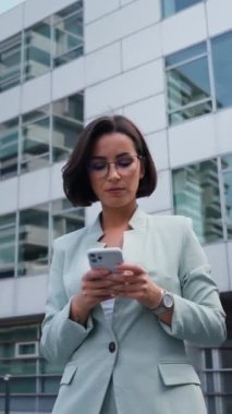 Young busy successful professional beautiful business woman holding cellphone and reading message while standing or walking on big city urban street outside during the break