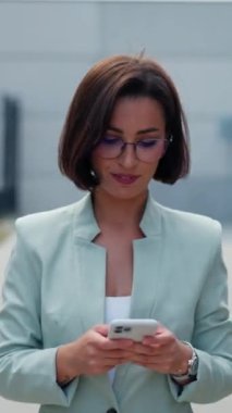 Charming positive young woman looking at the camera with smartphone on the city street. Businesswoman texting on the way to the office. Brunette adult girl messaging in social networks outdoors