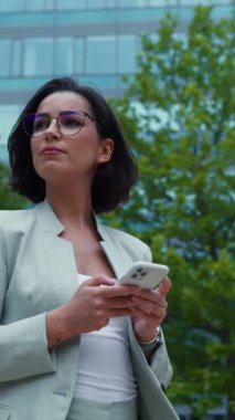 Serious businesswoman standing near office modern business center and writing message at her smartphone. Lady boss waiting for colleague