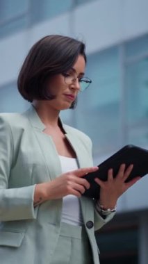 Vertical view of the beautiful young woman using tablet computer while walking through night city street full of lights. Portrait of gorgeous smiling female using her device