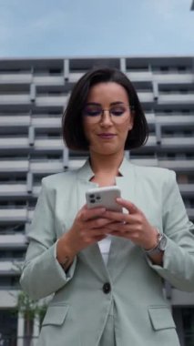 Female realtor wearing formal suit holding smartphone while going through the financial district. Businesswoman near the road in the urban space