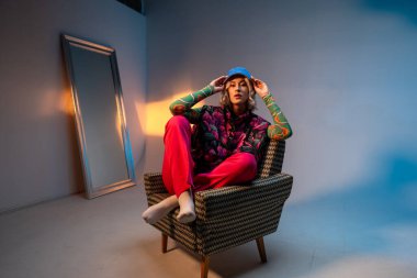 Havalı hippi öğrenci kadın şık giyinmiş kameraya bakıyor ve şapkasını düzeltiyor. Beyaz kadın model koltukta oturuyor.