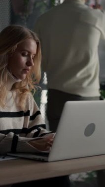 Blonde caucasian girl wearing casual clothes typing laptop computer in green plants coworking office. Her colleagues chatting at the background