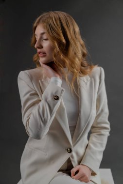 Bir portre zarif, dalgalı saçlı, şık beyaz takım elbiseli, taburede poz veren zarif bir kadını yakalar.