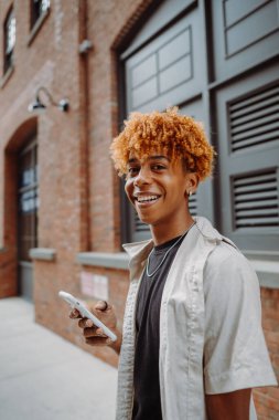 Kıvırcık saçlı, gülümseyen ve elinde akıllı bir telefon tutan genç bir adam..