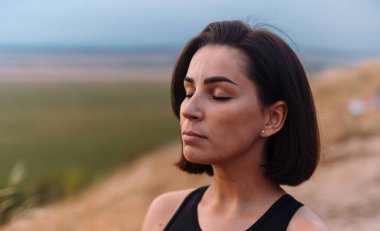 Bir kadın dışarıda huzur içinde meditasyon yapıyor, gözleri kapalı, gün batımında doğayı kucaklıyor, sakin manzarayla çevrili.