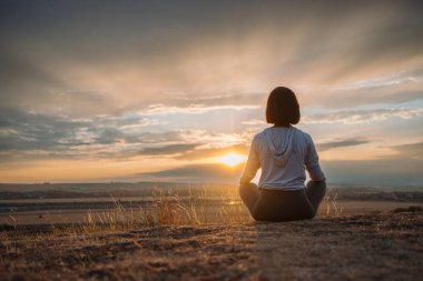 Gün batımında bir tepede huzur içinde meditasyon yapan bir birey, doğanın dinginliği ve güzelliğiyle harmanlanır.