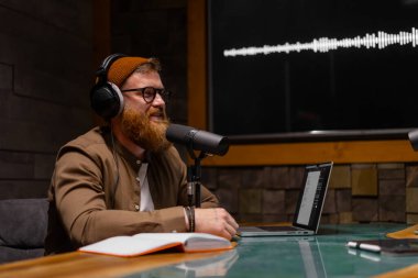 Sakallı bir adam yaratıcı içeriği için bir tablet ve mikrofon kullanarak bir stüdyoda podcast kaydediyor.