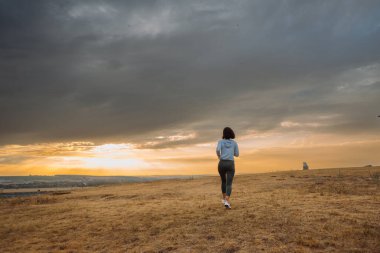 Gün batımında, sakin ve huzurlu bir ortamda dramatik bulutlarla çevrili, uçsuz bucaksız bir tarlada neşeyle koşan bir insan.