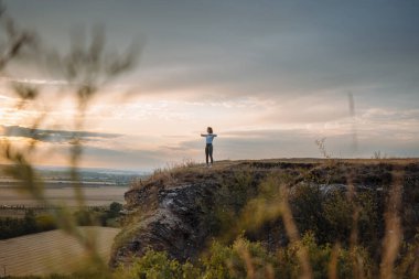 Huzurlu bir insan uçurumun kenarında, kolları açık, nefes kesici bir günbatımının tadını çıkarıyor, sükunet ve yalnızlığı hissediyor.