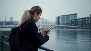 Kendine güvenen ve zarif bir kadın, Cola sonbahar havasında nehir kıyısında tek başına dururken gülümseyerek akıllı telefon yazıyor.