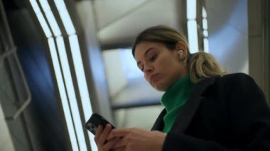 Akıllı telefonu kullanan iş kadını, arka planda metro istasyonunda yürüyen merdivende duruyor.