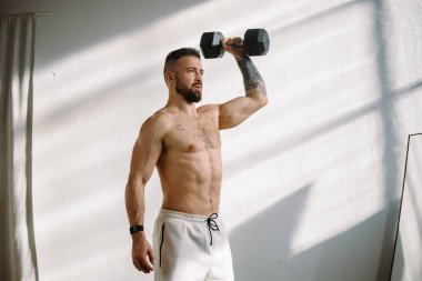 Parlak ve güneşli bir spor salonunda, kaslı bir adam yoğun bir şekilde bir halterle egzersiz yapıyor.
