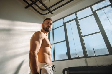 Kaslı bir adam, güneşli bir spor salonunda kendinden emin ve gururlu bir şekilde duruyor.