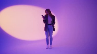 20 'li yaşlarda resmi ceketli genç bir kadın dansediyor ve neon rengi mor arka plan stüdyosunda izole bir şekilde poz veriyor. İnsanların yaşam tarzı konsepti. Dans ediyor, eğleniyor, jestler yapıyor.