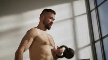 Tişörtsüz bir adam, titrek bir spor salonunda gücünü gösteren bir su ısıtıcısını kaldırır.