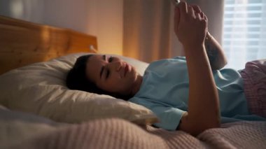 Genç bir birey, yatakta rahat bir şekilde yatarken akıllı telefonuna bakmaktan mutlu olur.