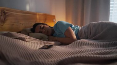 Bir kadın, sakin bir sabah sahnesinde yavaşça rahat yatağında uyanır ve telefonuna uzanır.