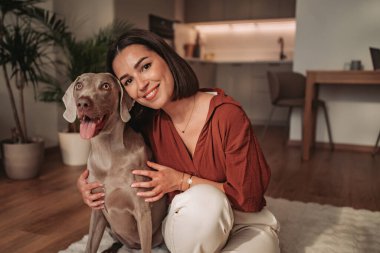 Neşeli bir kadın modern bir oturma odasında sevgili köpeğine sarılıyor.