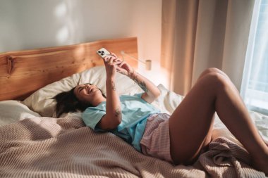 Neşeli bir kadın, huzurlu sabah rutininden zevk alır. Telefonunu yatakta kaydırırken.