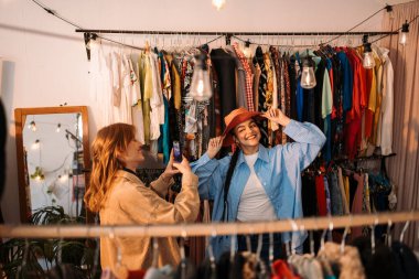 İki arkadaş, büyüleyici bir vintage mağazasında moda kıyafetleri denerken mutlu bir şekilde fotoğraf çekiyorlar.