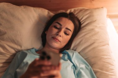 Bir kadın rahat bir şekilde yatakta uzanıyor, rahat bir gece atmosferinde akıllı telefonunu kullanıyor.