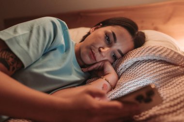 Genç bir kadın rahat bir şekilde yatağında uzanıyor, dikkatle akıllı telefonuna odaklanıyor.