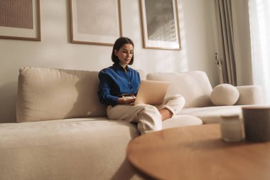 Odaklanmış bir kadın laptopunu parlak, modern bir yaşam alanında koltukta kullanır.