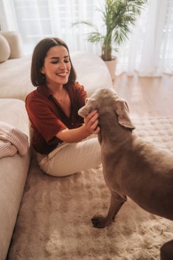 Bir kadın, sıcak ve sevgi dolu bir mekanda köpeğiyle neşe içinde oynuyor ve bağ kuruyor.
