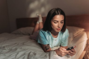 Parlak, güneşli, rahat bir yatak odasında, genç bir kadın telefonunu çeşitli görevler için mutlulukla kullanıyor.