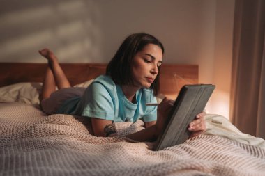Bir kadın rahat bir şekilde yatağında uzanıyor, tabletini sıcacık bir ortamda tutuyor.
