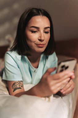 Genç bir kadın rahat bir yatakta uzanırken akıllı telefonunu kullanarak harika vakit geçiriyor.