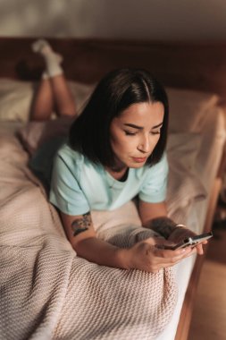 Bir kadın rahat bir yatak odasında dinlenirken akıllı telefonuyla mutlu mesut eğleniyor.