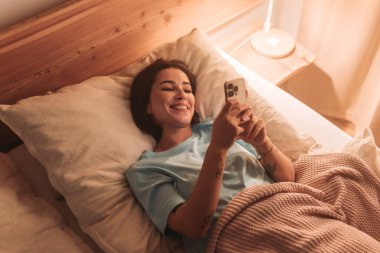 Bir kadın yatakta uzanırken telefonuna gülümsüyor, boş zamanlarının tadını çıkarıyor..