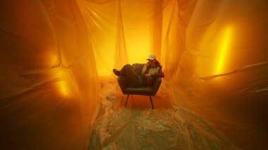 Yalnız bir figür, gerçeküstü sarı bir arka planla çevrili eski bir sandalyede rahatça oturur.