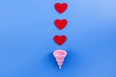 Silikon pembe regl fincanı düz. Kadın sağlığı ve alternatif hijyen. İçinde kan damlaları ya da mavi arka planda kırmızı kalpler olan bir fincan. Çevre dostu, kadın hijyeni için alternatif yeniden kullanılabilir ürün..
