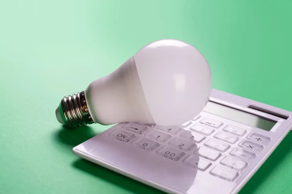 White calculator and LED bulb on green background. Concept showing the payment of electricity bills. The concept of savings electricity. Reducing the payment of utility bills